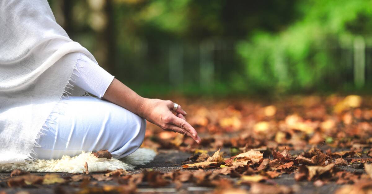 A woman sitting outside among fallen leaves, hand poised on her knee as she meditates.