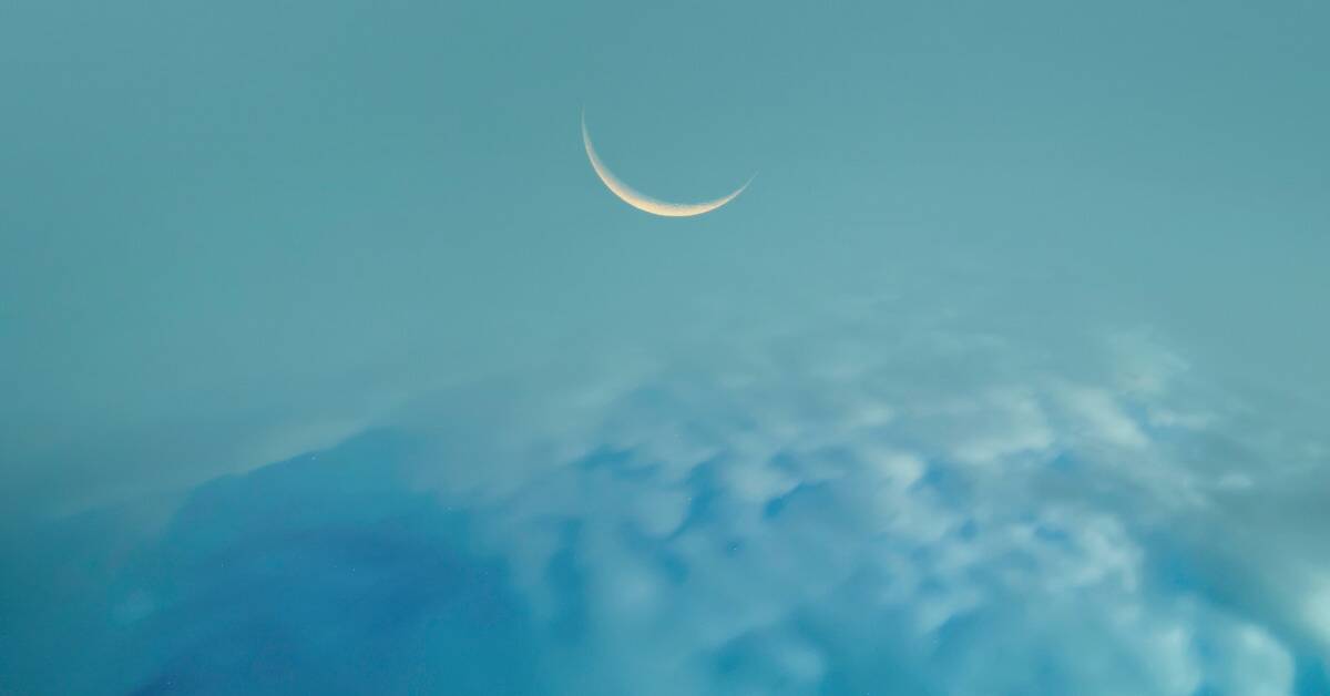 A thin new moon in a vibrant blue sky above some clouds.
