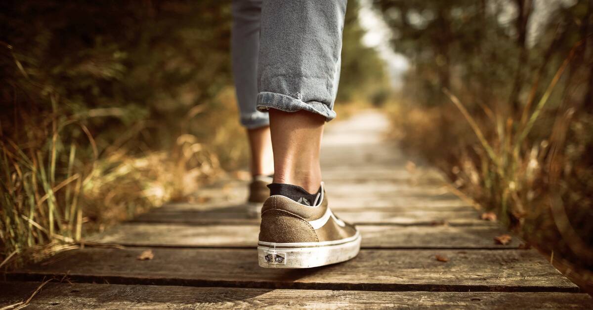 A close shot of someone in capris and skate shoes walking down a wooden boardwalk with tall grass on either side.