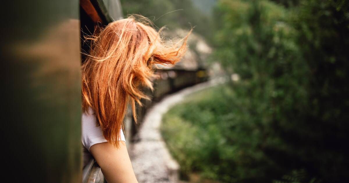 A woman with bright red hair leaning out of a train window as it rides the track, hair blowing in the wind.