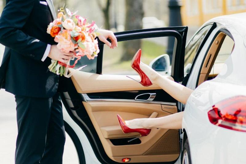 A man standing in front of an open car door with a bouquet of flowers. A woman's legs sporting red high heels are sticking out from that car door.