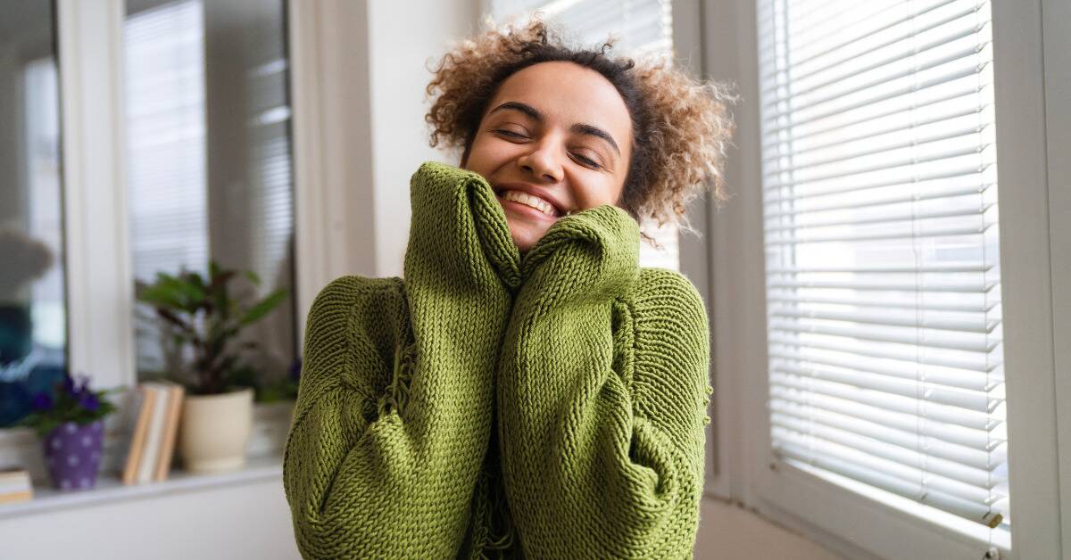 A woman in a large green sweater smiling as she rests her chin in her sleeve-covered hands.