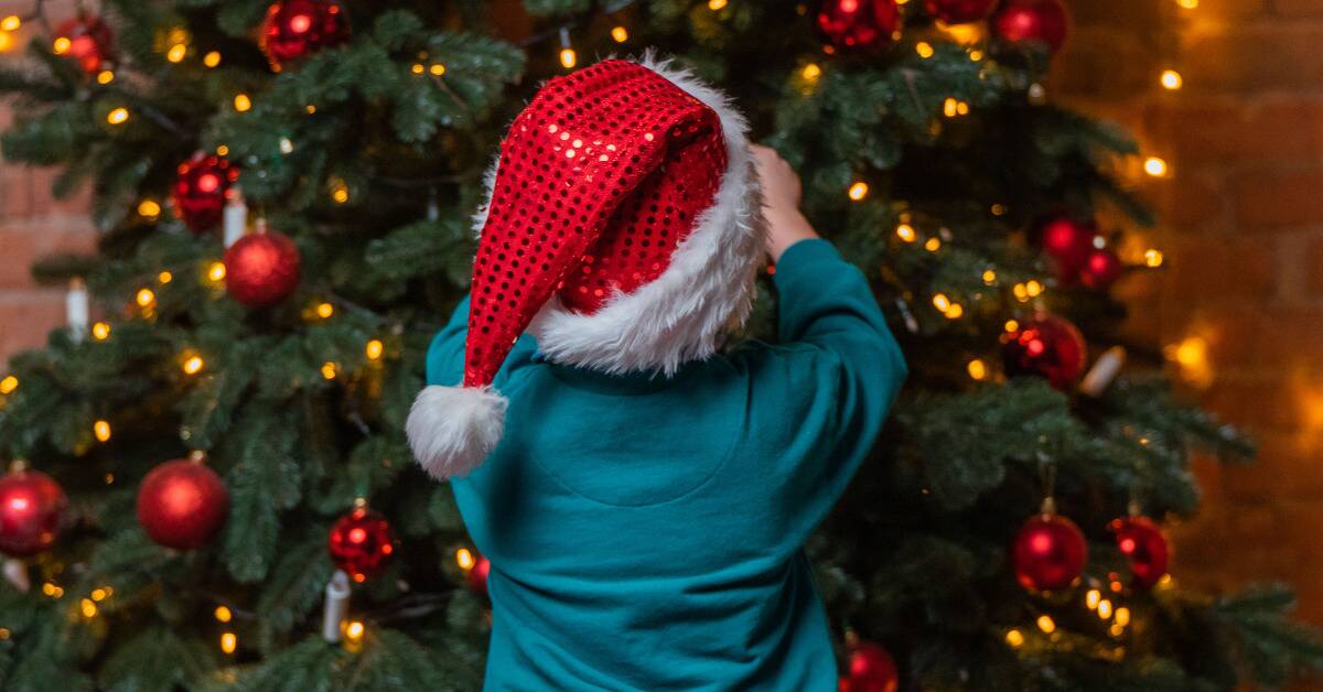 A small child in a sparkly red Santa hat putting an ornament on a Christmas tree.