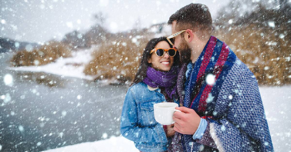 A couple standing outside as it snows, smiling at one another, one holding a warm mug.
