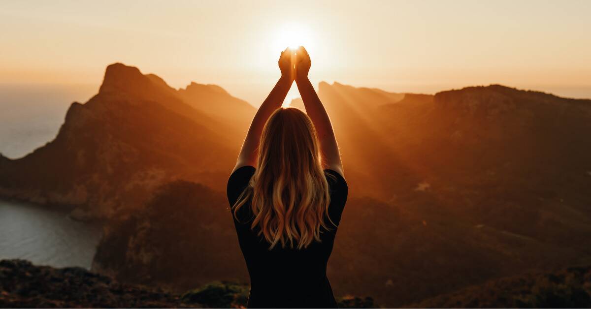 A woman facing a mountain range at a sunrise, holding her hands up so it appears she's cupping the sun in them.