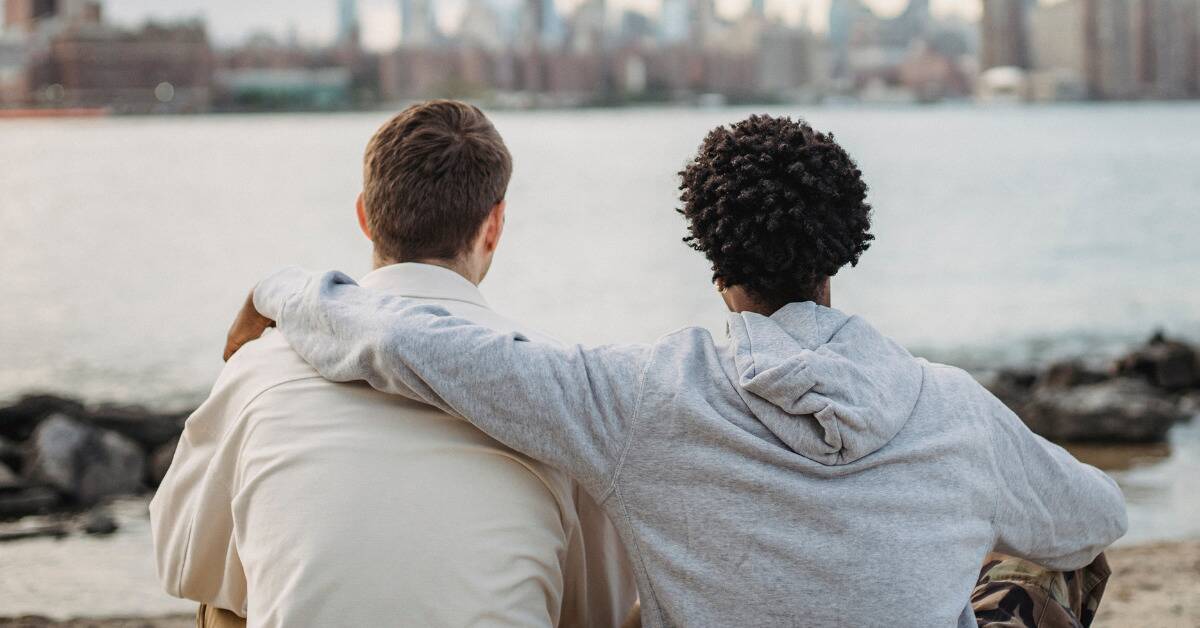 Two friends sitting in front of the water, one with his arm around the other in a comforting gesture.