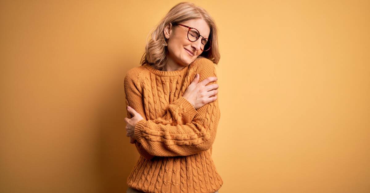 A woman in a yellow sweater against a yellow background, wrapping her arms around herself, hugging herself as she smiles with her eyes closed.