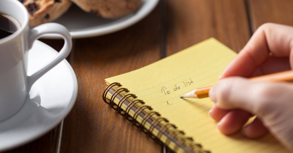 Someone writing their to-do list in a yellow-paged notebook on a table next to a coffee mug and a plate with some cookies.