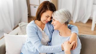 A mother and her adult daughter sitting on a couch, hugging close and looking into each other's eyes.
