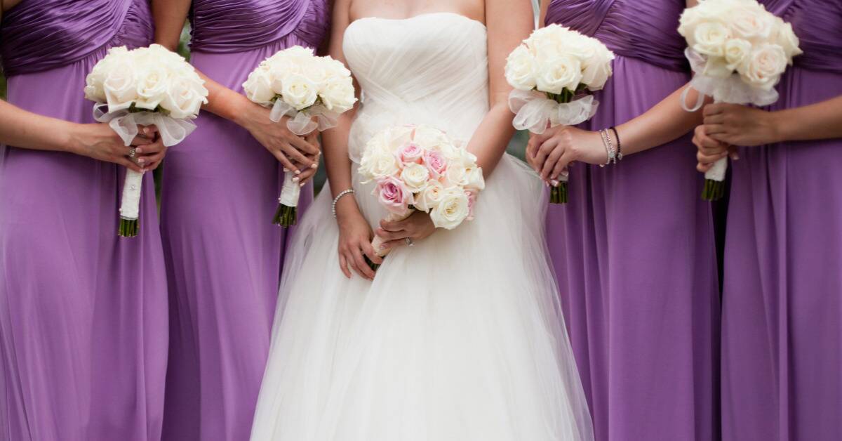 A bride standing in the middle of her bridesmaids, who are in purple dresses, everyone holding their bouquet.