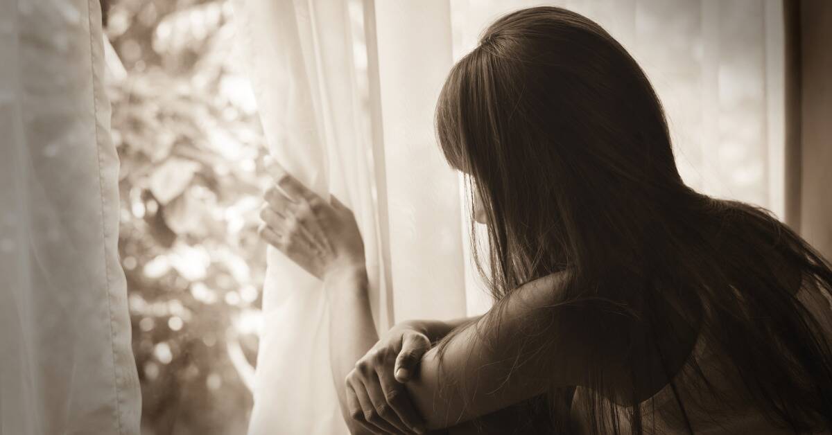 A muted image of a woman looking out a window, gently drawing the curtain aside as her hair hides her face.