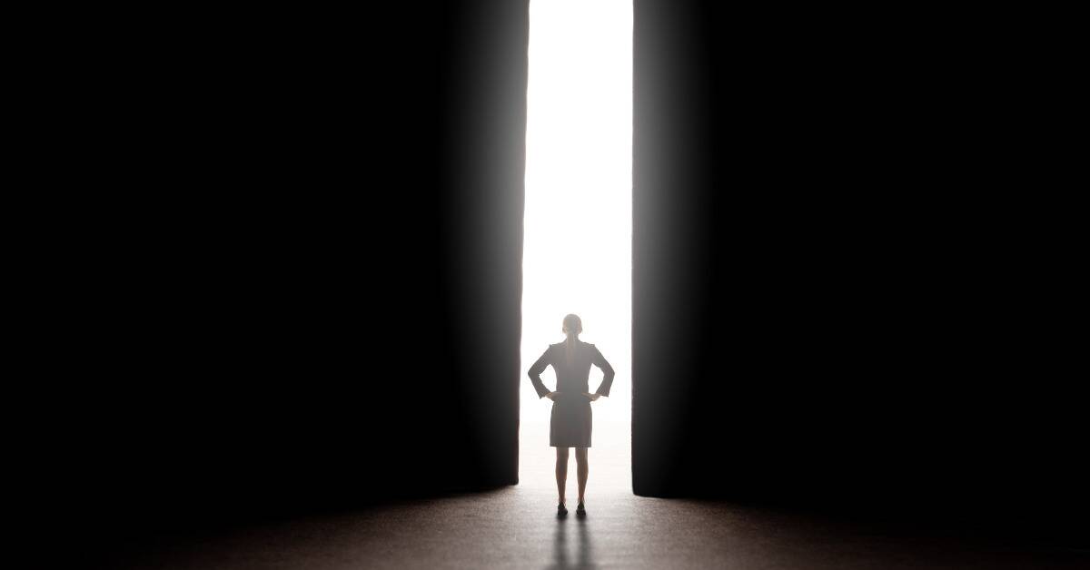 A businesswoman standing in front of a large pillar of light between two large, dark doors that are opening up to her.