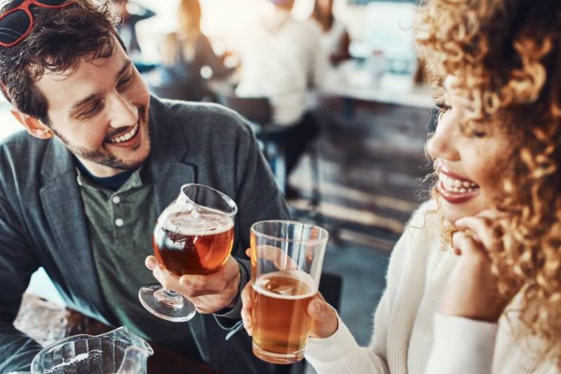 Two people meeting at a bar, both smiling and clinking their glasses of beer together.
