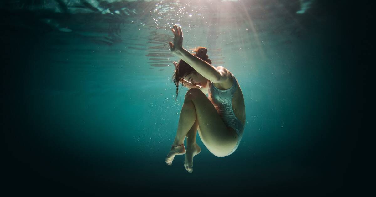 A woman falling underwater gracefully, legs drawn to her chest as she floats down.
