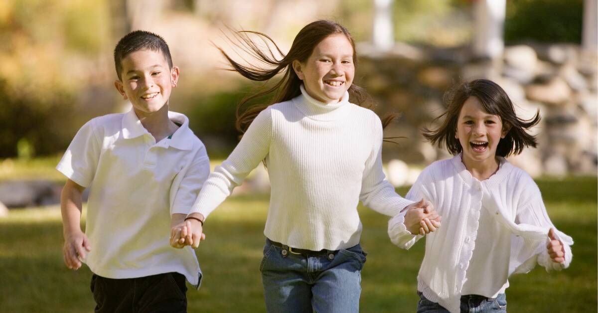Three young siblings all smiling as they run outside, all holding hands.