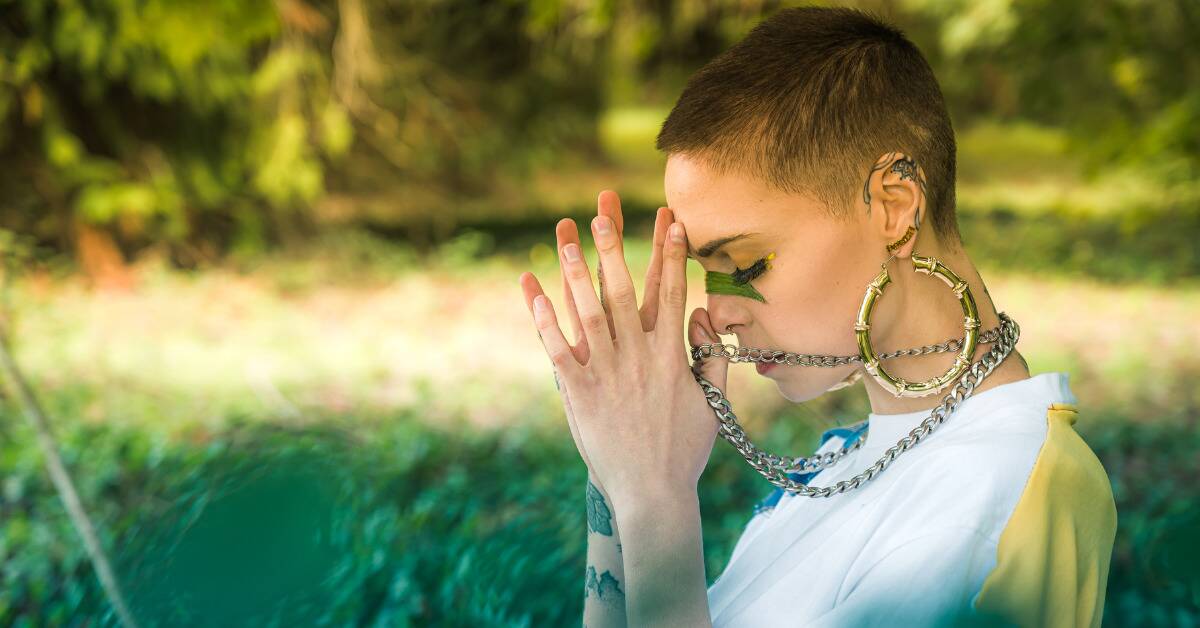 A woman in bold jewelry standing outside, eyes closed and hands together by her face, seeming to be in the middle of a prayer.
