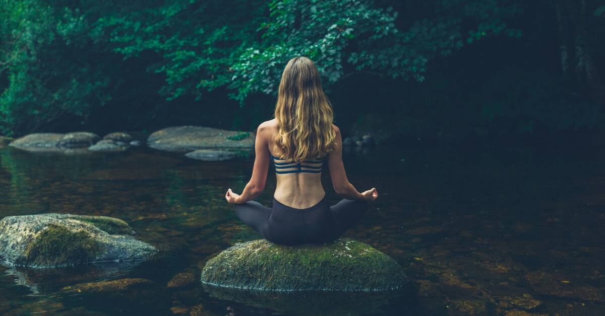 A woman sitting on a mossy rock in the middle of a creek, legs folded as s he meditates.