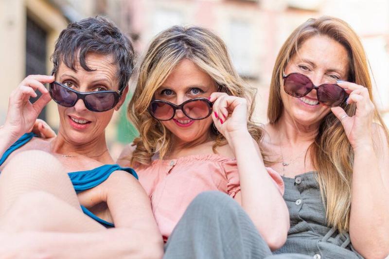 Three freinds tilting down their sunglasses to look at the camera, all smiling or laughing.