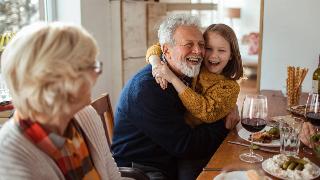 A granddaughter hugging her grandfather at a dinner table while her grandmother watches.