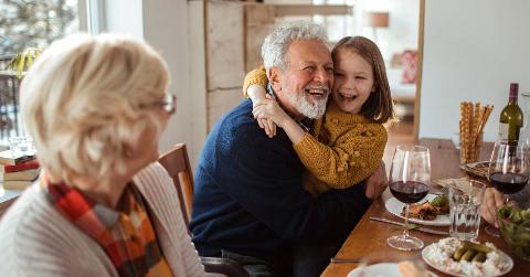 A granddaughter hugging her grandfather at a dinner table while her grandmother watches.