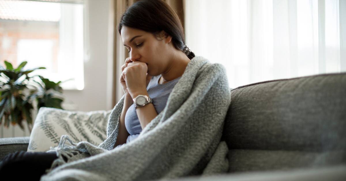 A woman sitting on her couch with a blanket wrapped around her, a fist to her mouth as if she's mid-cough.