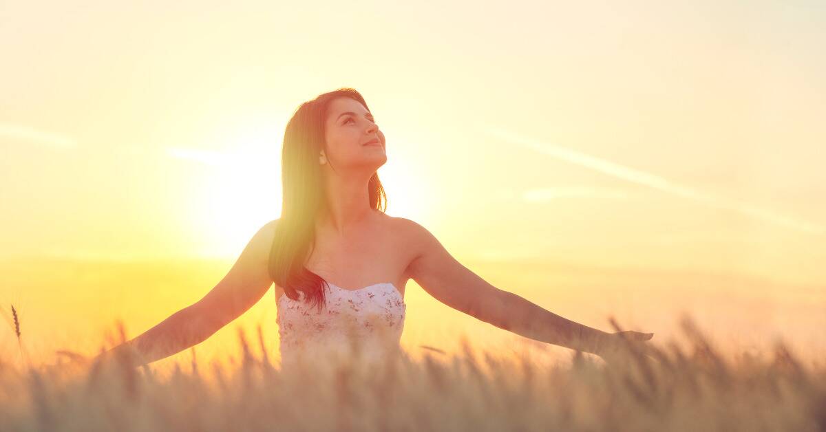 A woman walking through a field of tall grass, her arms out, looking up at the sky with a smile as the sun rises behind her.