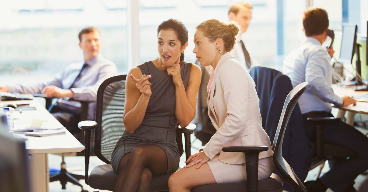 Two women in an office setting gossiping, one of them pointing at something they're both staring at.