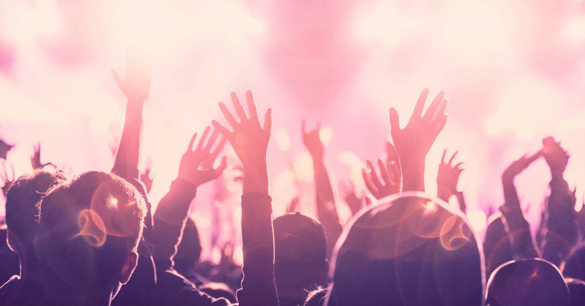 A crowd at a concert, lit buy bright pink lights, hands in the air as they cheer for the performer.