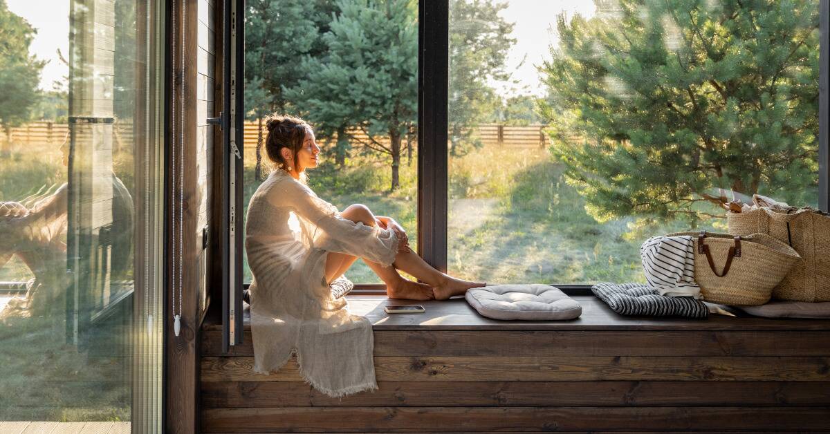 A woman sitting on a bench windowsill, smiling peacefully as she sun shines in.