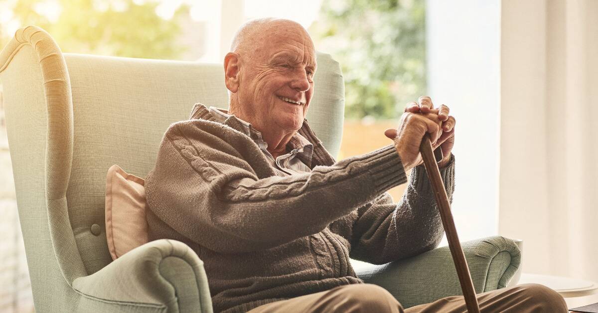 A senior man smiling while sitting in a chair by a large window.