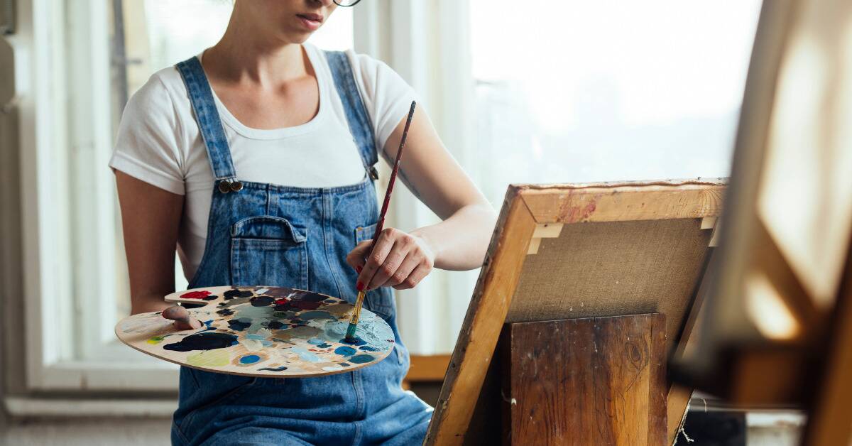 A woman sat at a canvas, holding a paint pallet, dipping her brush in some paint.