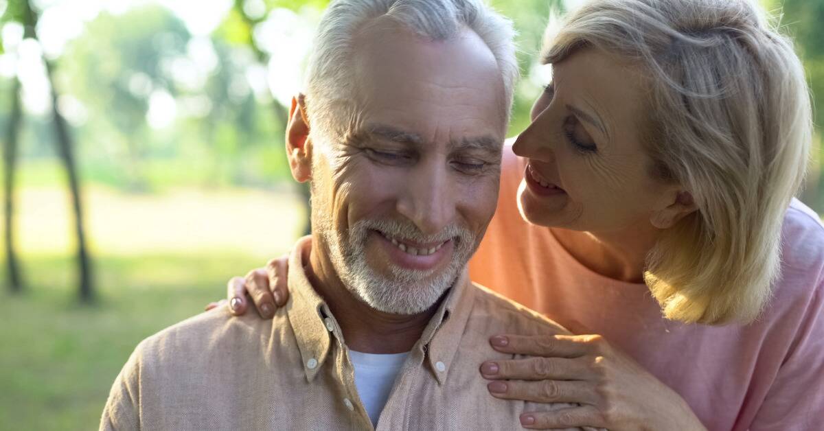 A woman leaning in very close to speak into a man's ear, her hands on his shoulders.