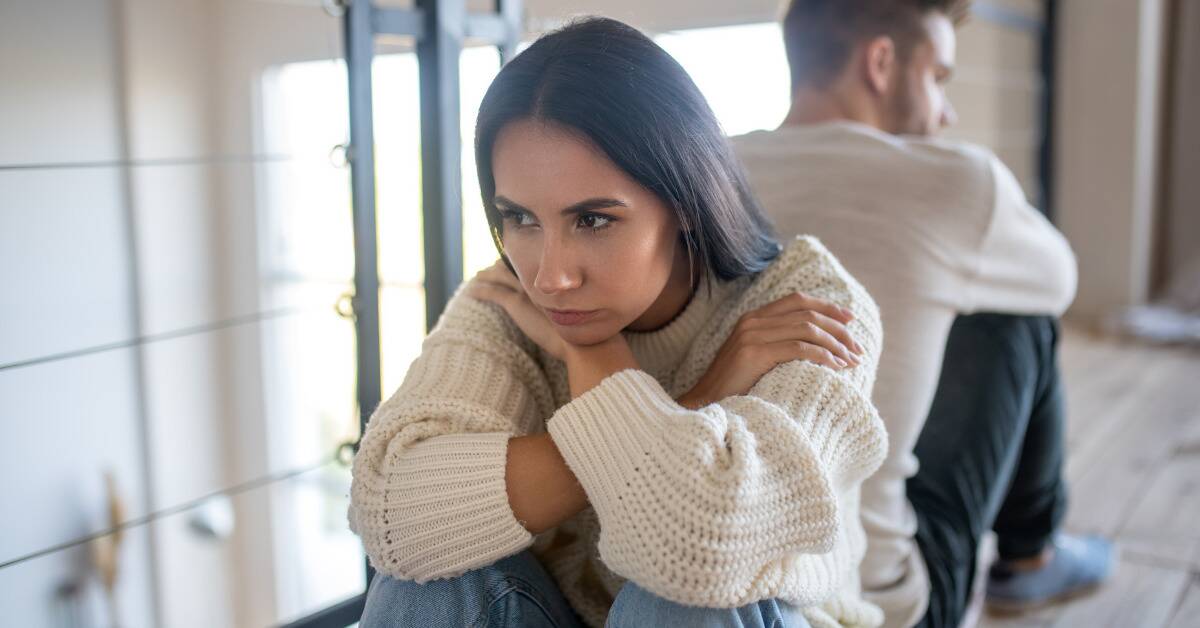 A woman sits on the floor, knees to her chest and arms crossed, looking upset and annoyed. Behind her, a man sits with his back to her, but it looking over his shoulder at her.