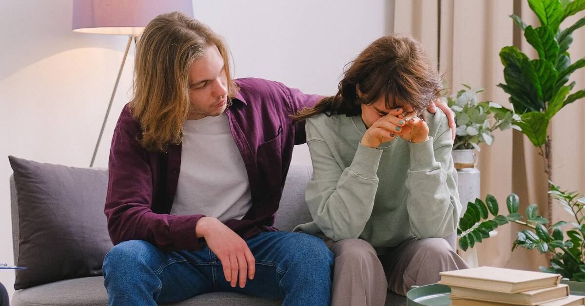 A couple sits on a couch, one half crying with her face in her hands while the other half puts a supportive arm around her.