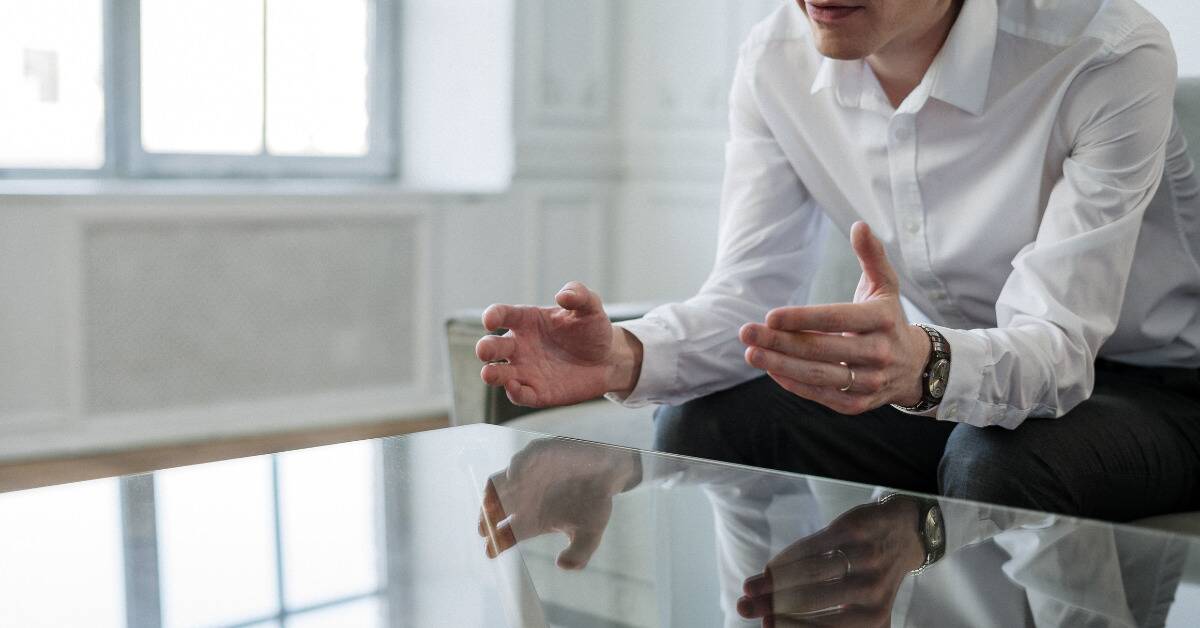 A man leaning forward, elbows on his knees, as he explains something to someone across a table.