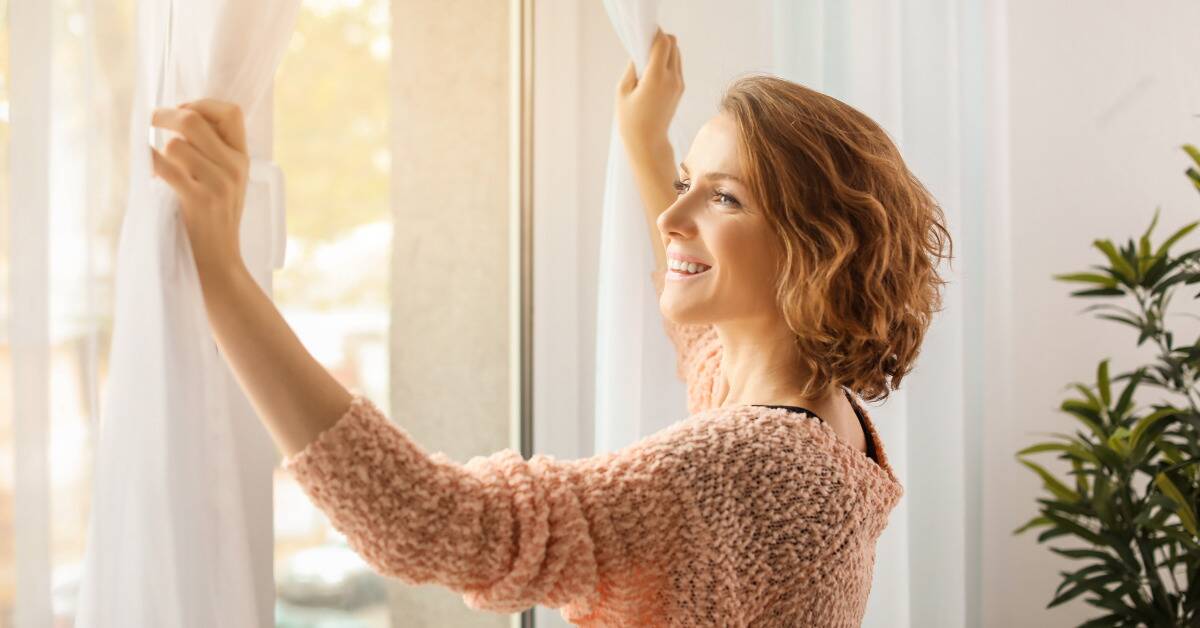 A woman opening curtains with a smile, letting sunlight shine into the room.