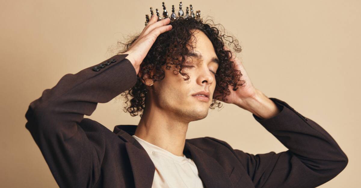 A man with his eyes closed lowering a black gem tiara onto his head.