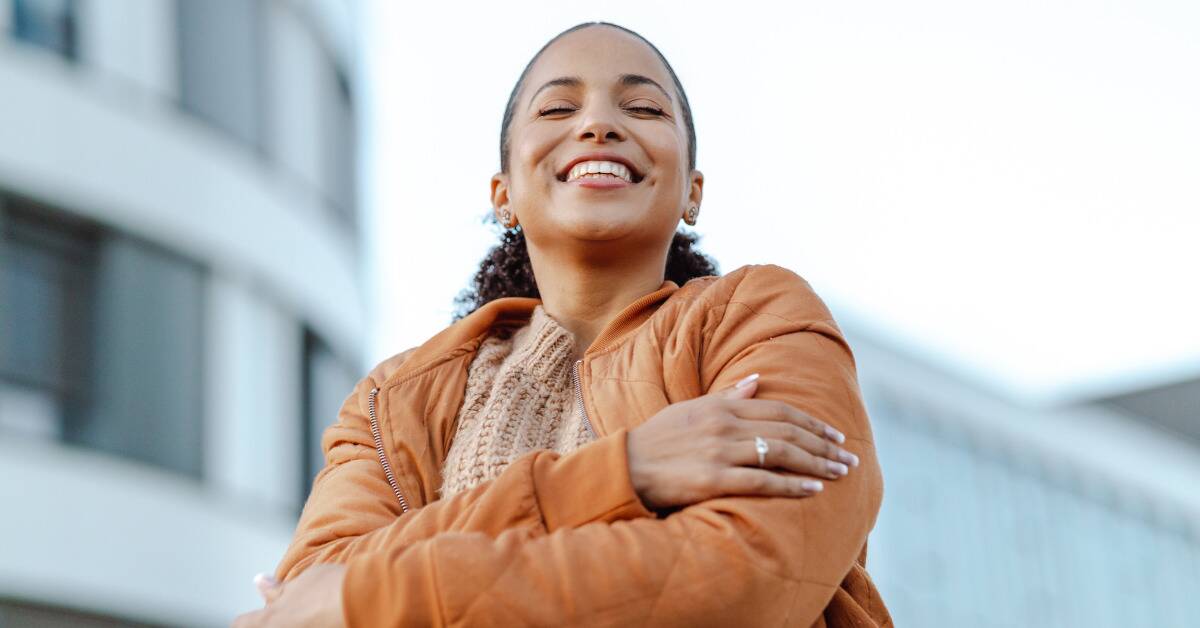 A woman standing outside, eyes closed and smiling, hugging herself.