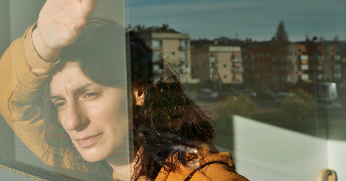 A woman leaning a hand and her forehead against a window, looking out at the world with a pained expression.