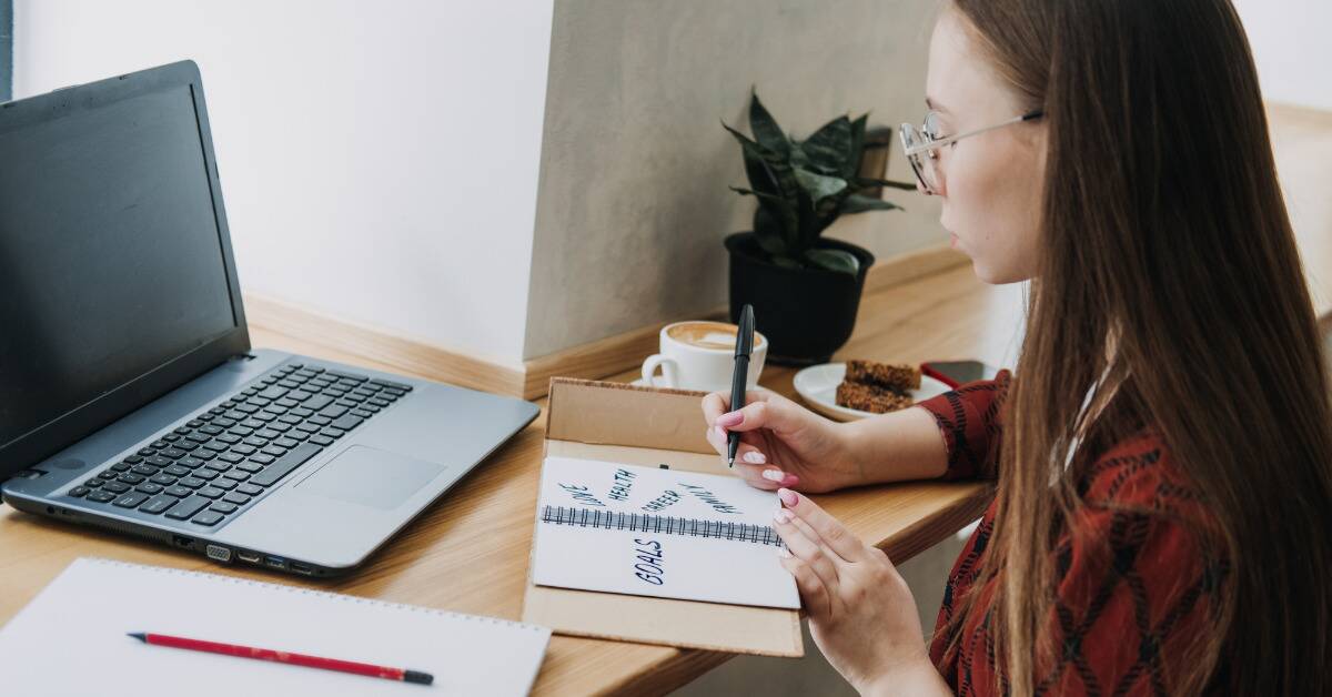 A woman is sat at her laptop, holding open a notebook with the world 'goals' written on it.