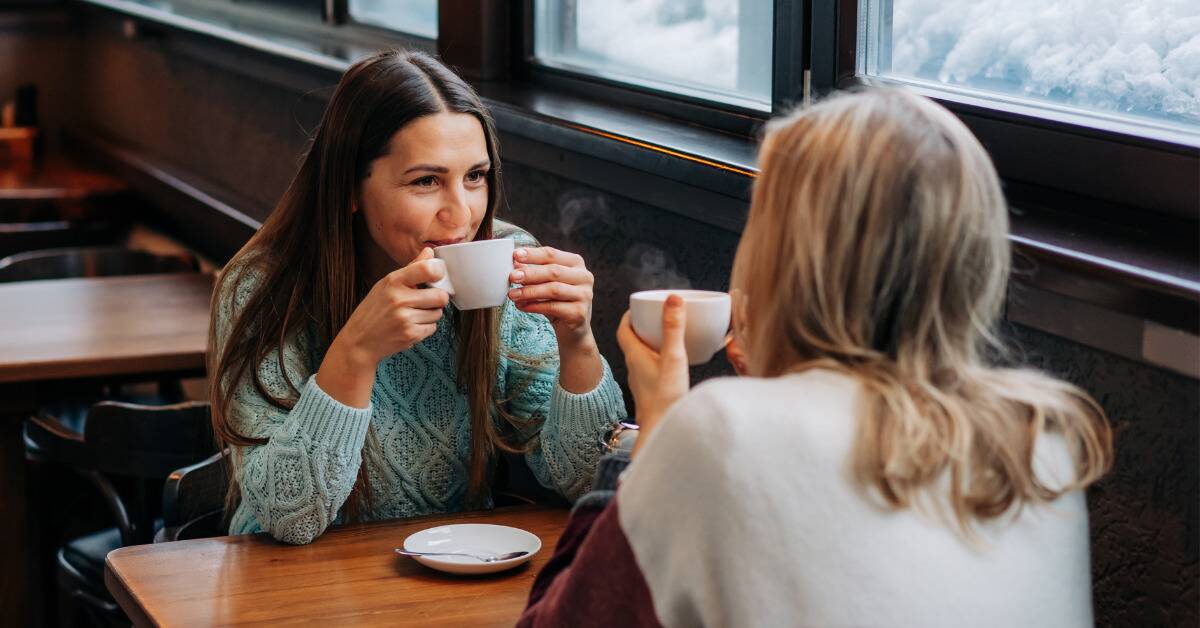 Two women sitting across from each other at a cafe, each holding mugs, smiling as they chat.