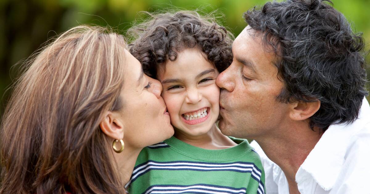 A mother and father each kissing one of their son's cheeks, the son smiling brightly between them.