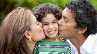 A mother and father each kissing one of their son's cheeks, the son smiling brightly between them.