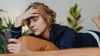 A woman anxiously looking at her phone as she lays on her stomach, hand on her forehead.
