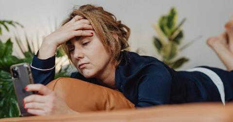 A woman anxiously looking at her phone as she lays on her stomach, hand on her forehead.