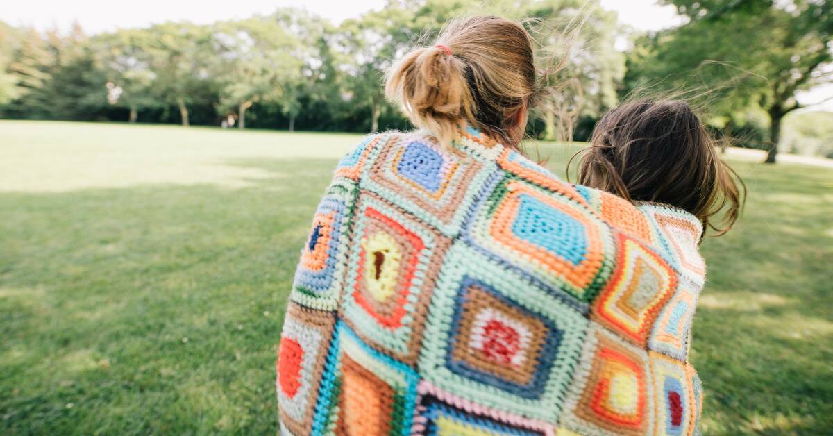 Two friends standing outside in the grass, a colorful blanket wrapped around both of their shoulders.