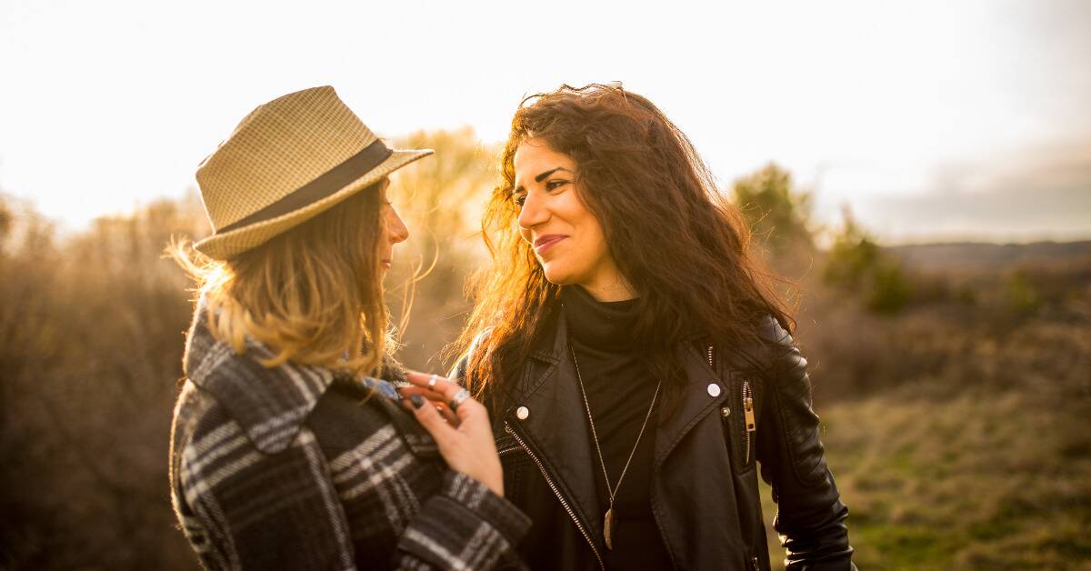 Two friends standing side by side outside in the sun, smiling at each other.