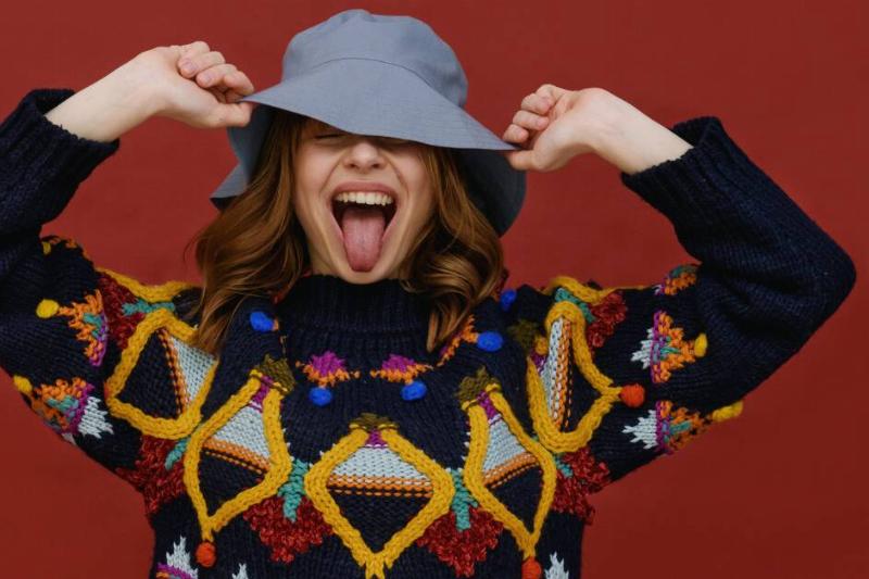A woman in a bright, busy sweater and a grey bucket hat, holding onto the brim of the hat as she sticks her tongue out.