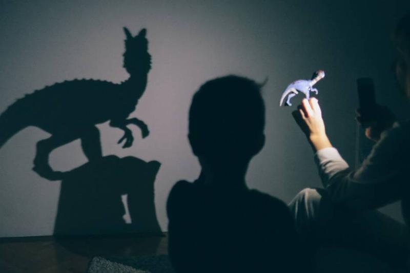 A mother using her phone's flashlight to project the shadow of a dinosaur toy onto the wall for her young son.