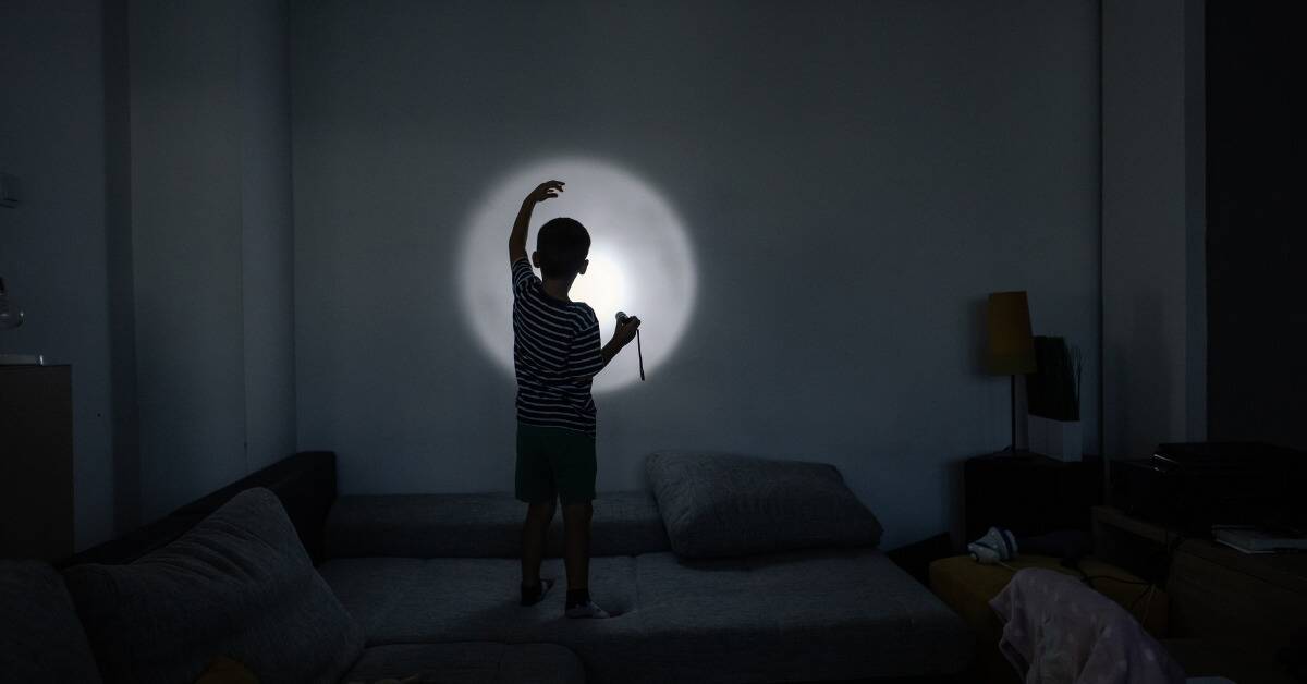 A young boy standing on a couch, holding a flashlight that he's projecting on a wall in a dark room, making shadow puppets.